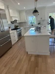 Bright white kitchen with island, stainless appliances, and a person standing by the window.