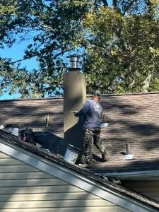 Person on a roof painting a chimney beside trees under a blue sky