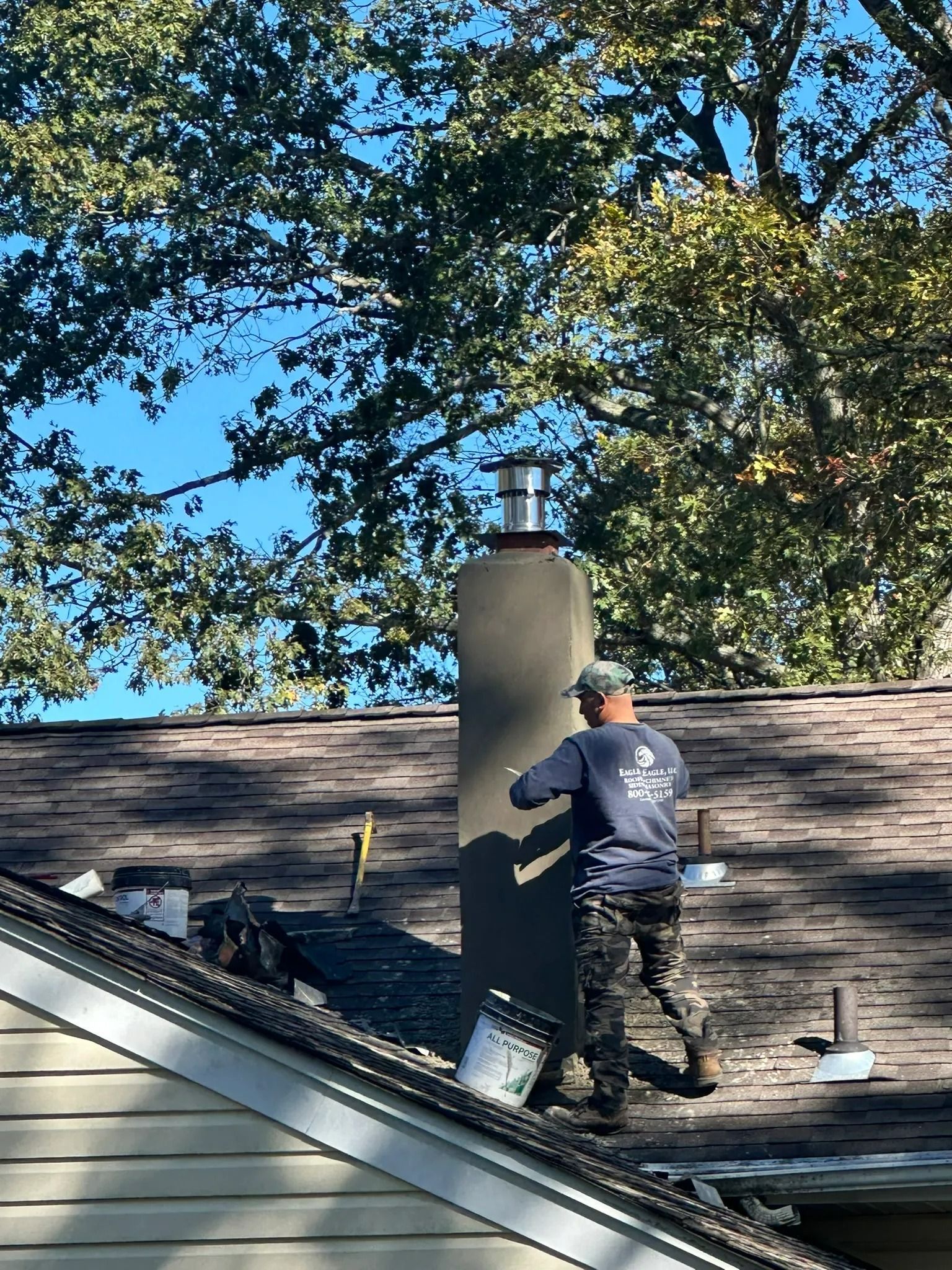 Worker on a shingled roof installing a tall chimney vent on a house under a blue sky