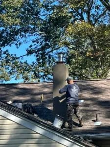 Worker on a roof inspecting a dark chimney beside trees and a blue sky.