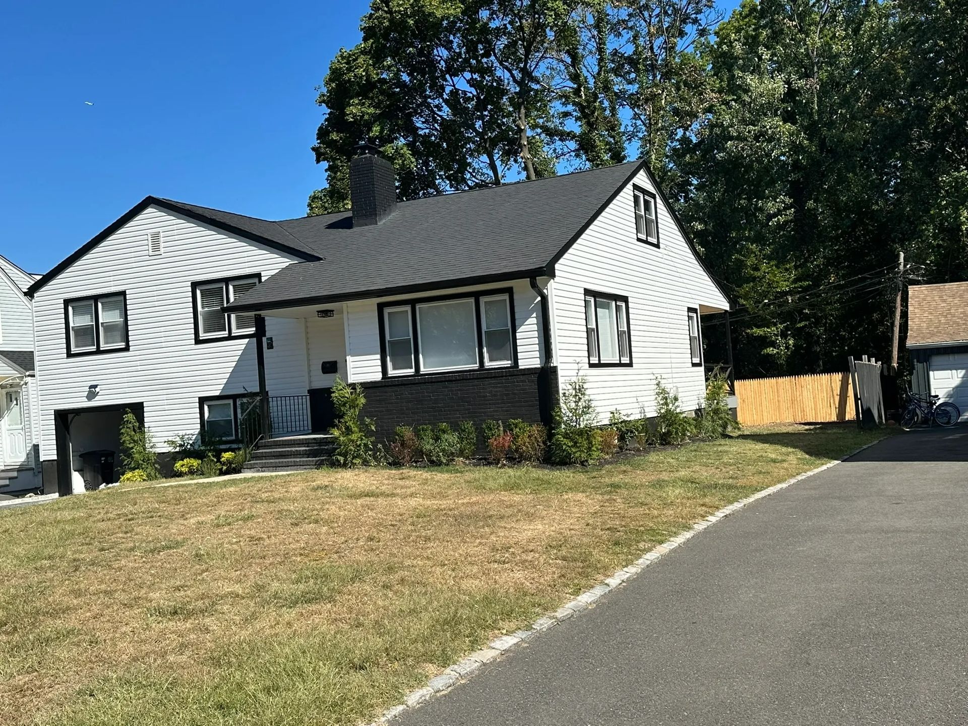 White house with gray roof and porch on a grassy slope, beside another house under a clear blue sky