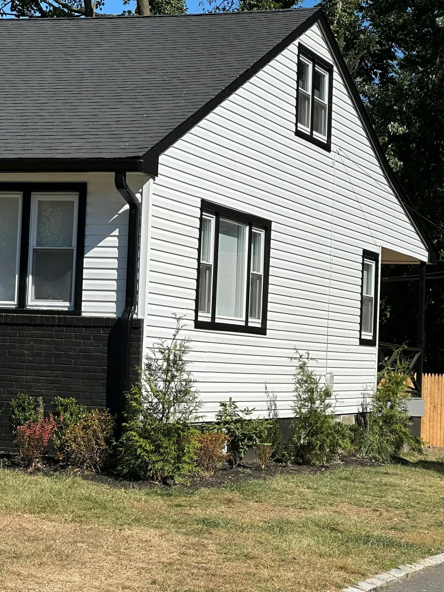White house side wall with gray roof, small windows, and shrubs in front under a sunny sky