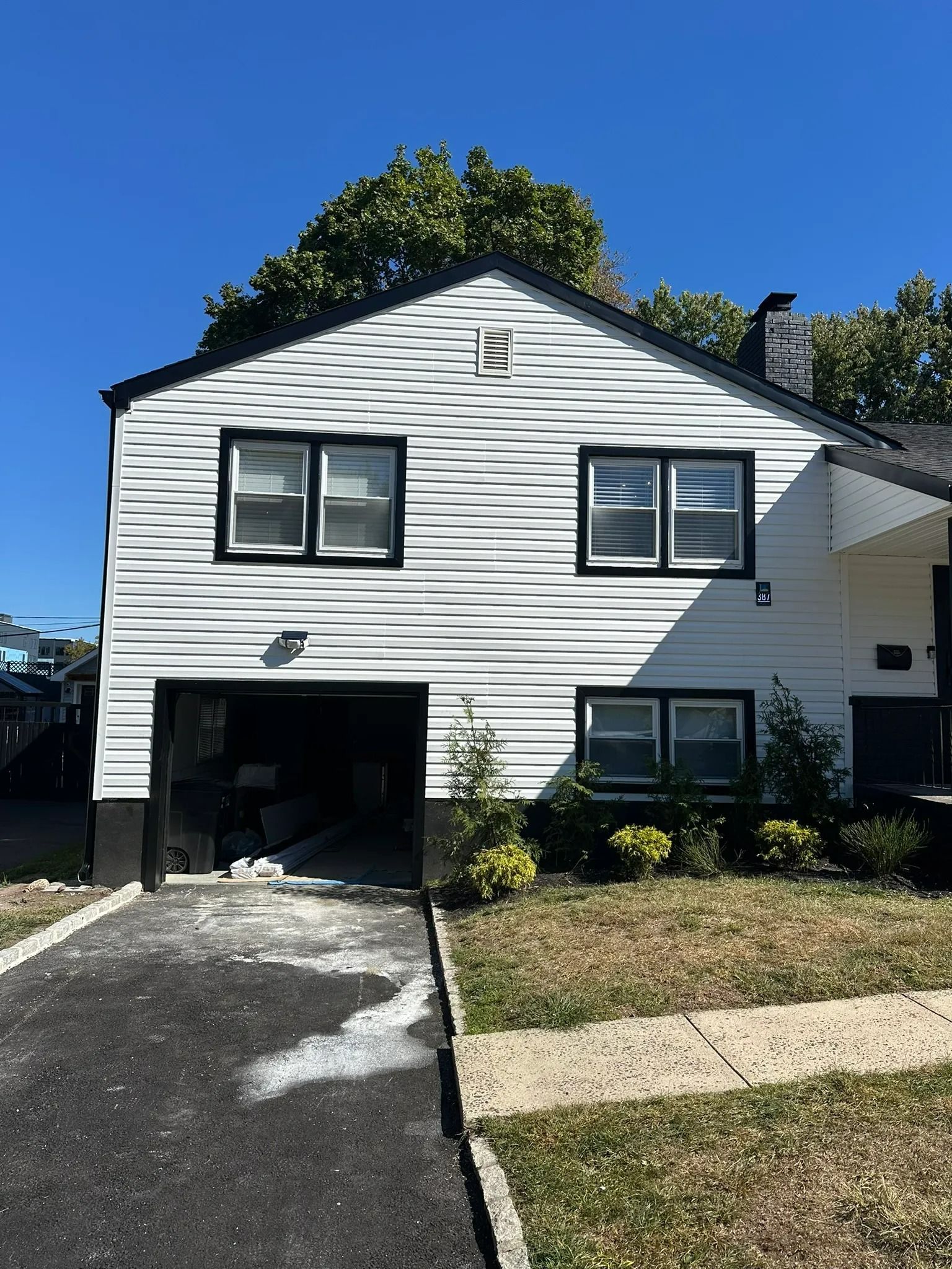 White two-story house with black shutters, garage opening, driveway, and blue sky.