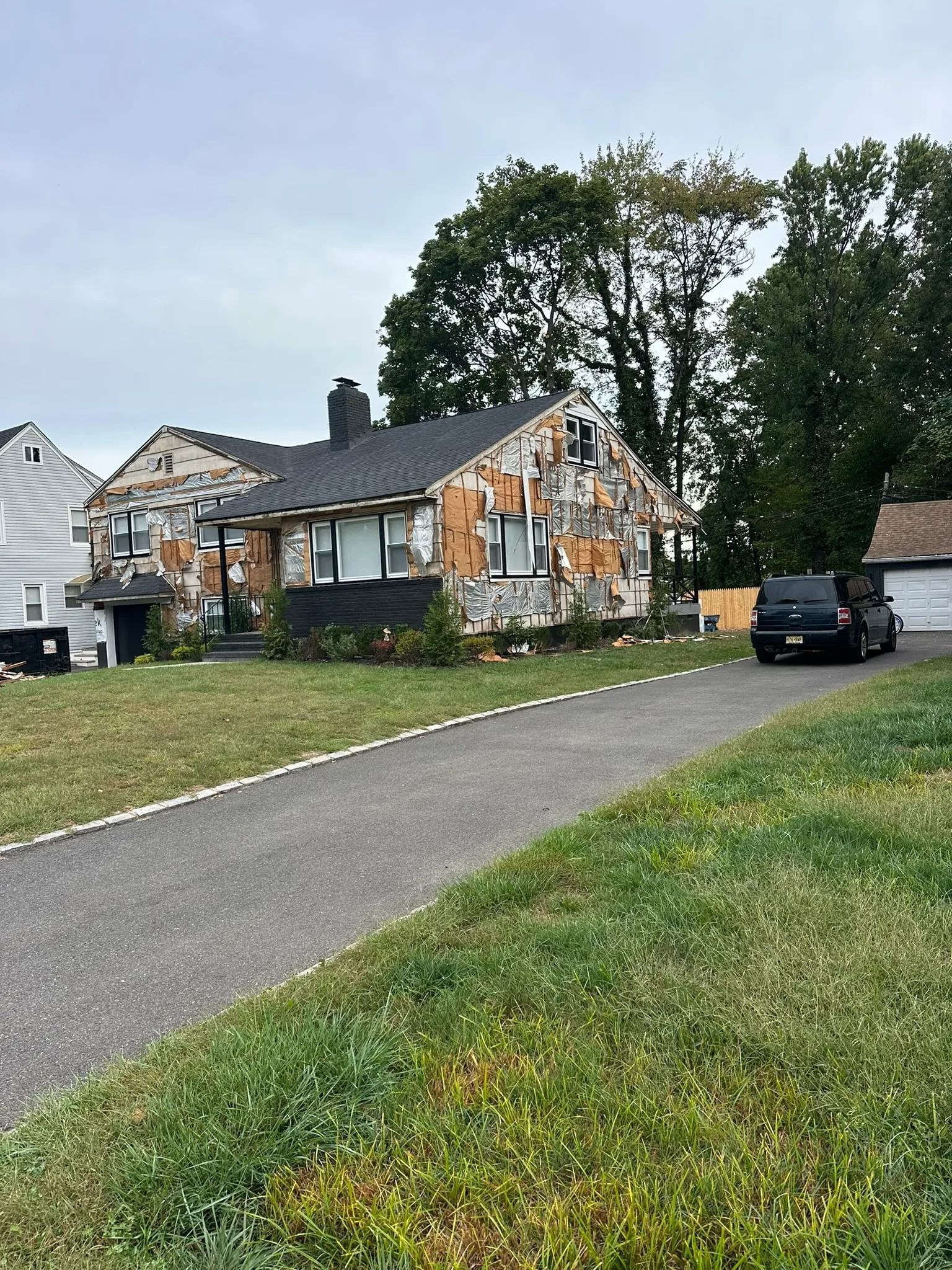 Suburban house with peeling exterior paint, driveway, parked SUV, and trees under an overcast sky
