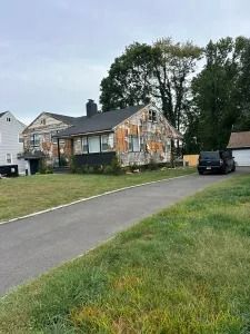Residential house with damaged siding, a driveway, parked black SUV, and tall trees in a grassy yard