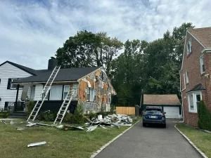 Partially demolished house with two ladders and debris in the front yard, next to a blue car in the driveway