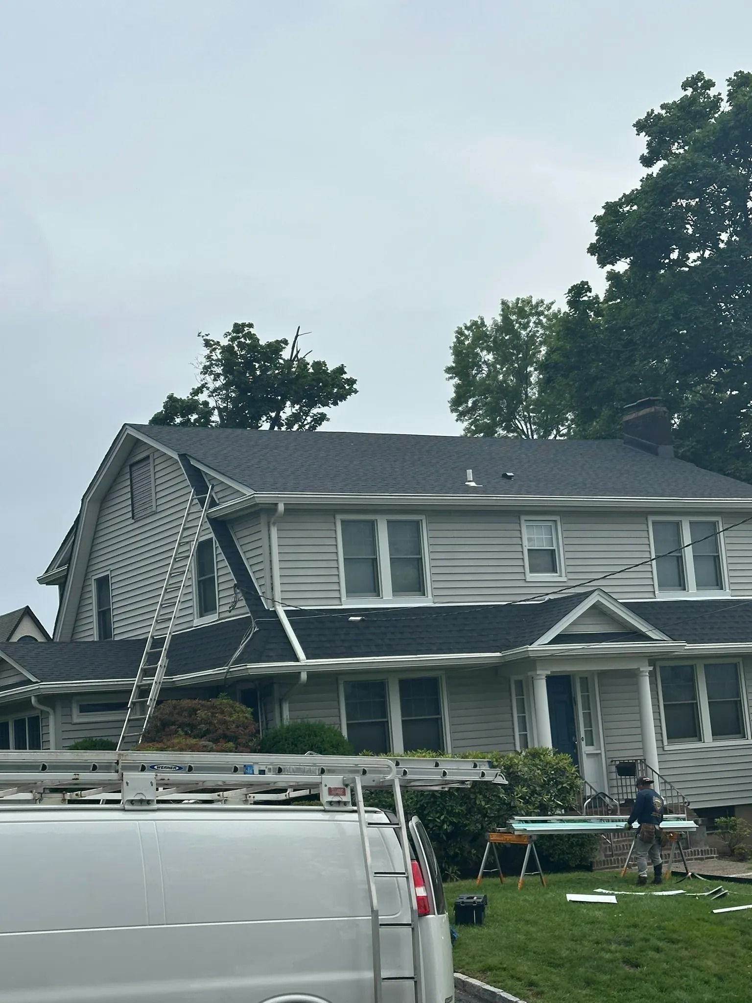 Damaged gray house with porch collapse behind a white van and scattered debris in the yard
