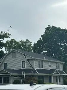 Gray two-story house with white trim and a porch, partially covered by a ladder, under a cloudy sky.
