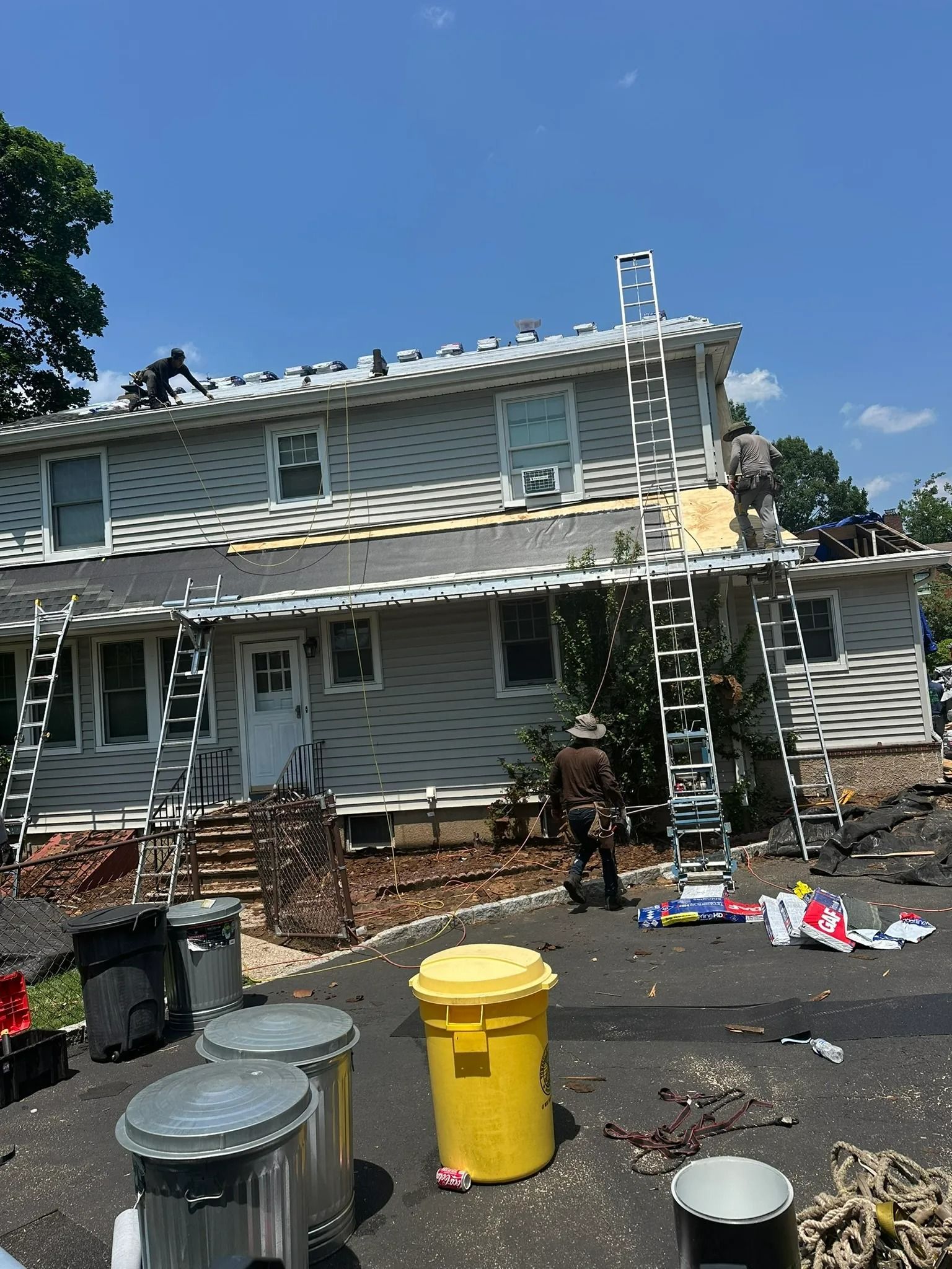 Damaged two-story house with fire damage, ladders, debris, and trash bins in the yard