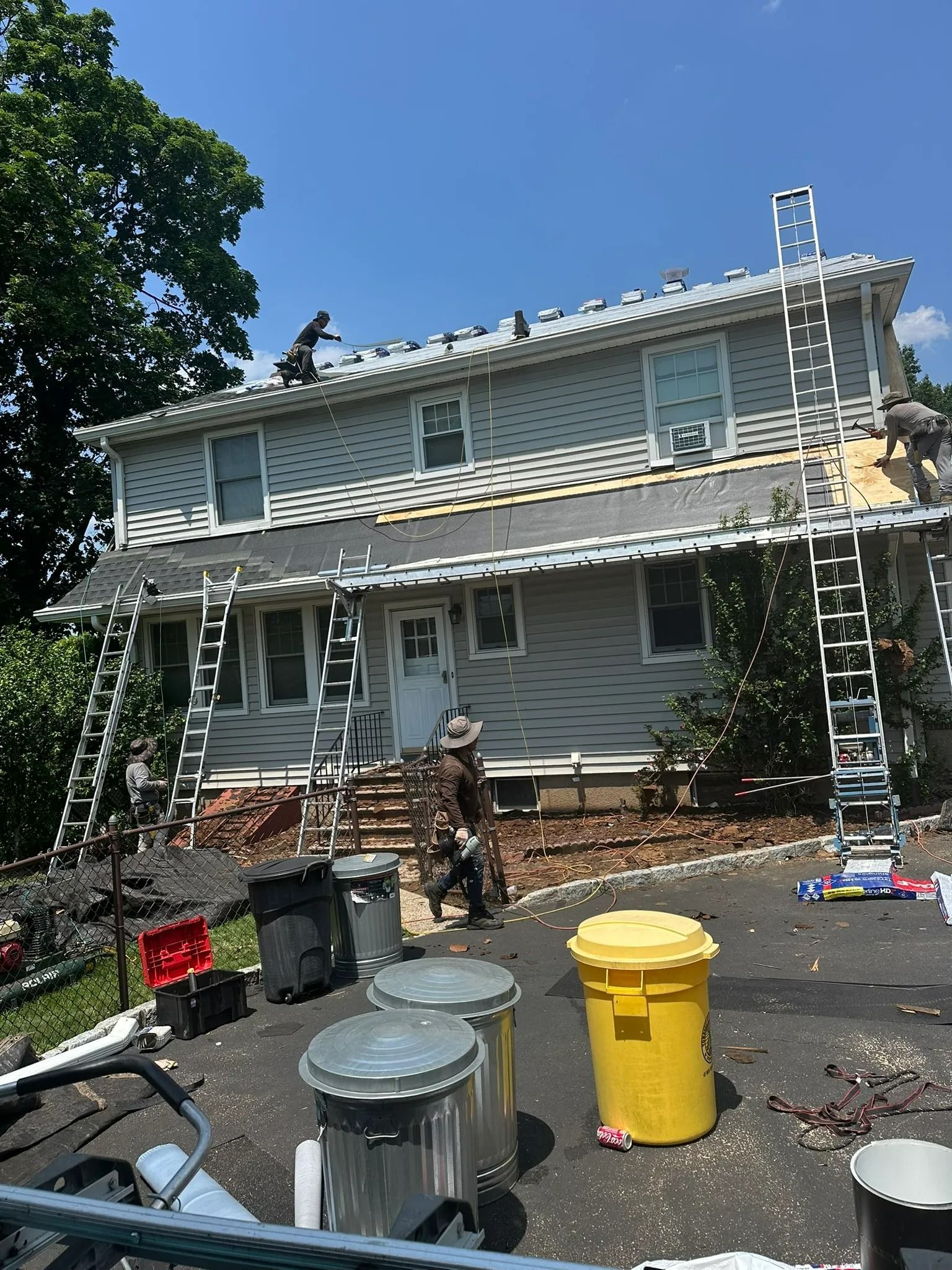 Two-story house with workers on ladders and trash bins in front, roof repair underway