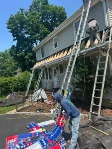 Worker on a ladder installing striped awnings on a house porch, with roofing supplies below.
