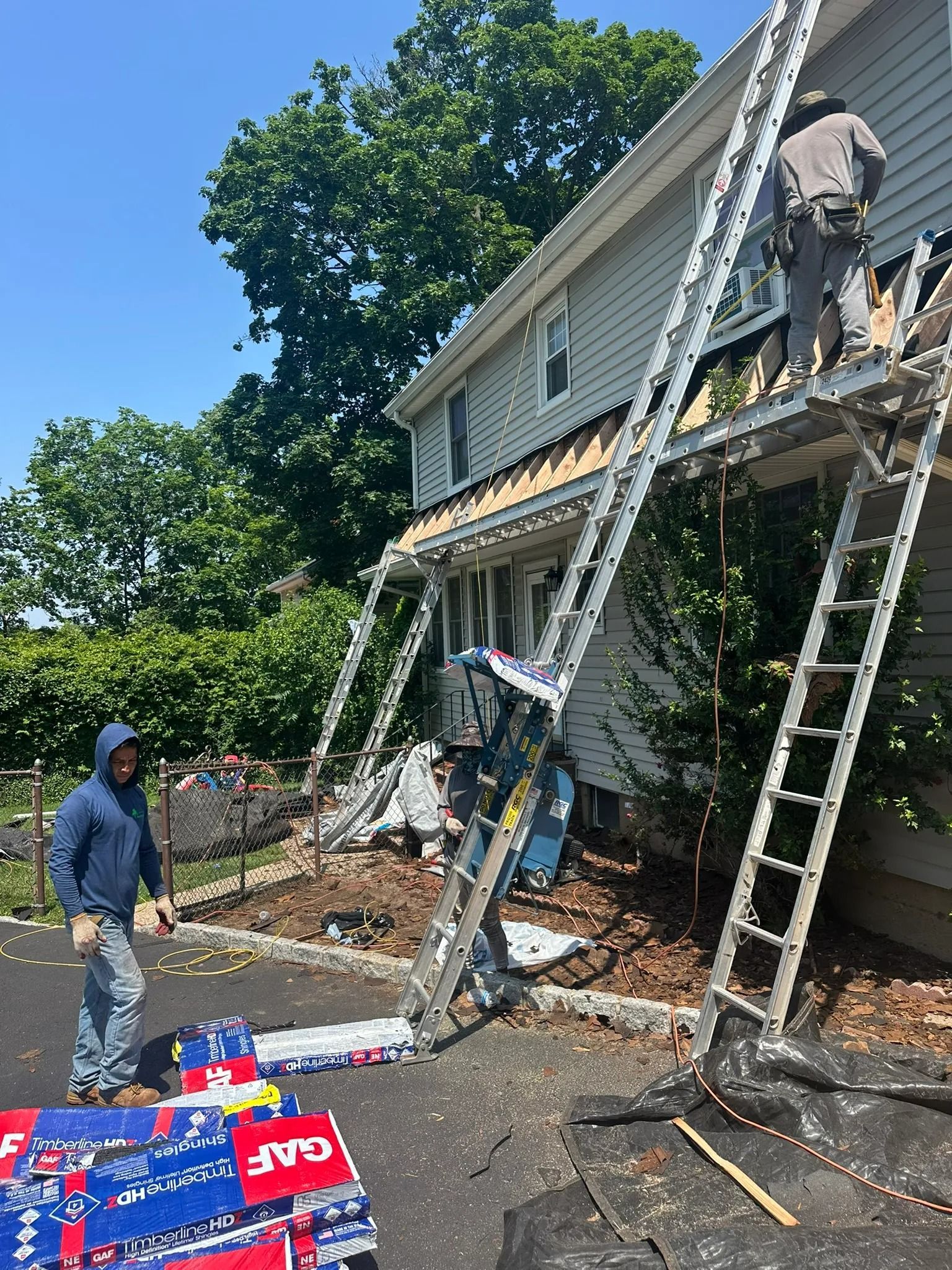 Workers on ladders repair a house roof and siding beside a pile of fallen debris.