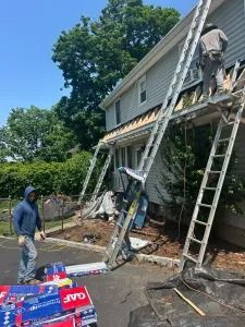 Workers on ladders repairing a house roof and siding beside scattered construction materials