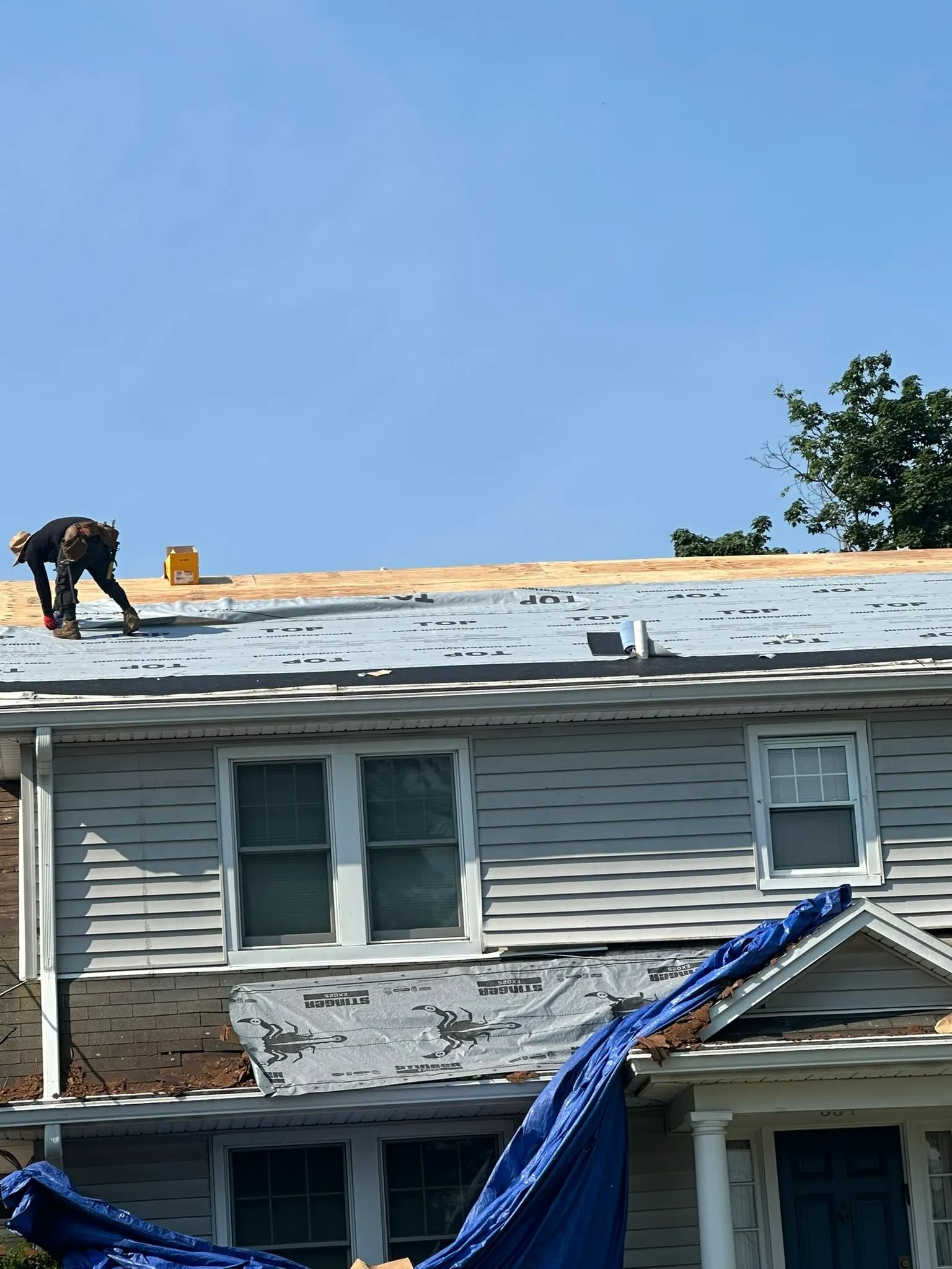 Roofing worker on a damaged house roof with blue tarp draped over the front wall