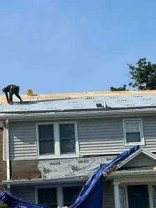 Worker on a roof repairing storm damage above a gray two-story house with a blue tarp draped over the front.