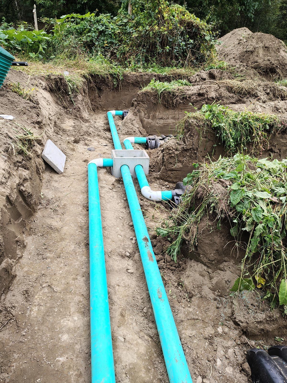 Blue pipes in a trench, leading to a gray junction box, outdoors in a hillside.