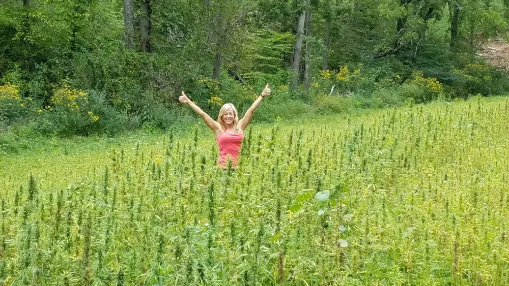 Franny Tacy stands in an outdoor hemp field that grows to her arm pits smiling and giving two thumbs way up