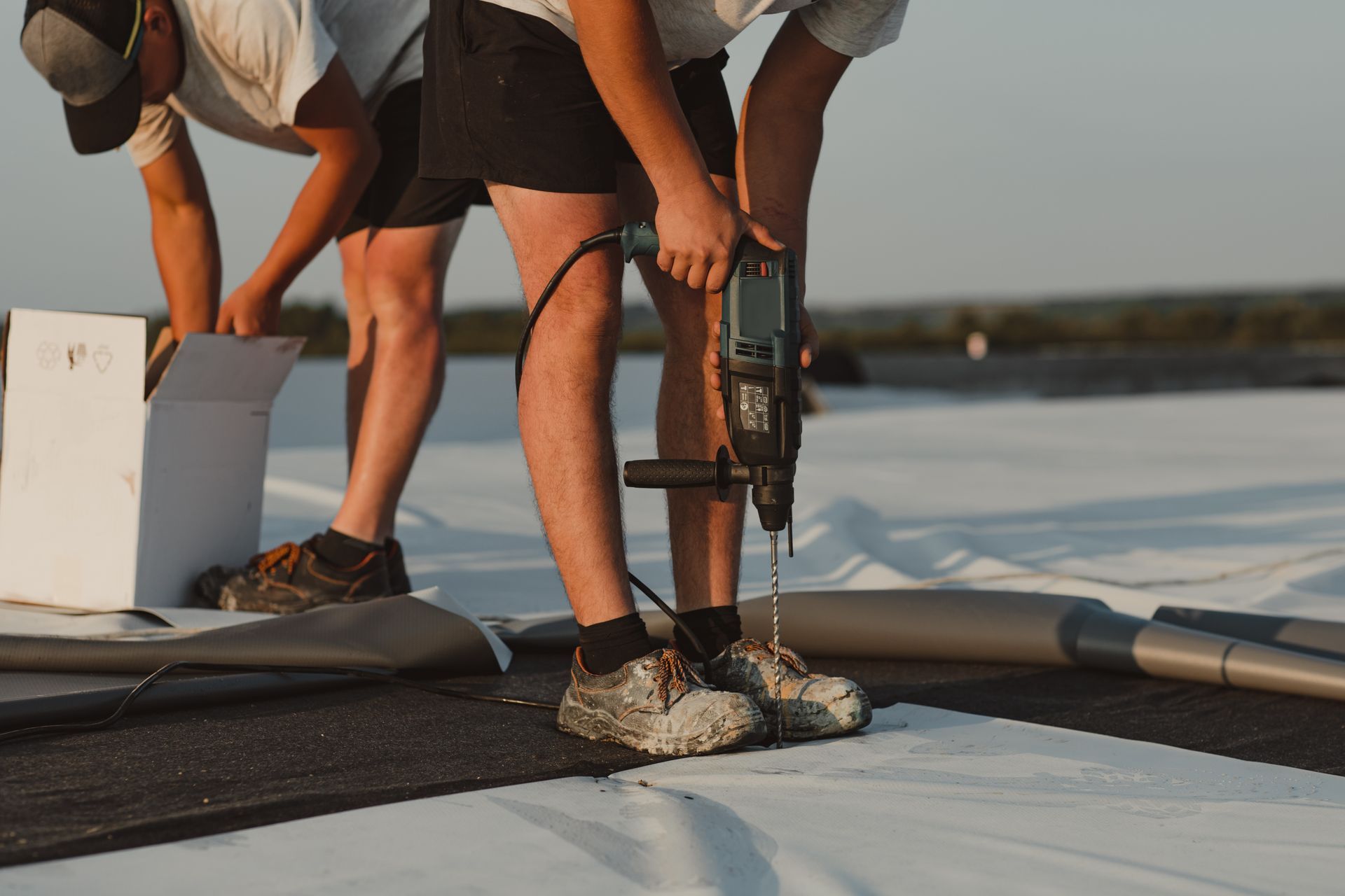 Workers install material on a commercial roof as one drills into the surface.