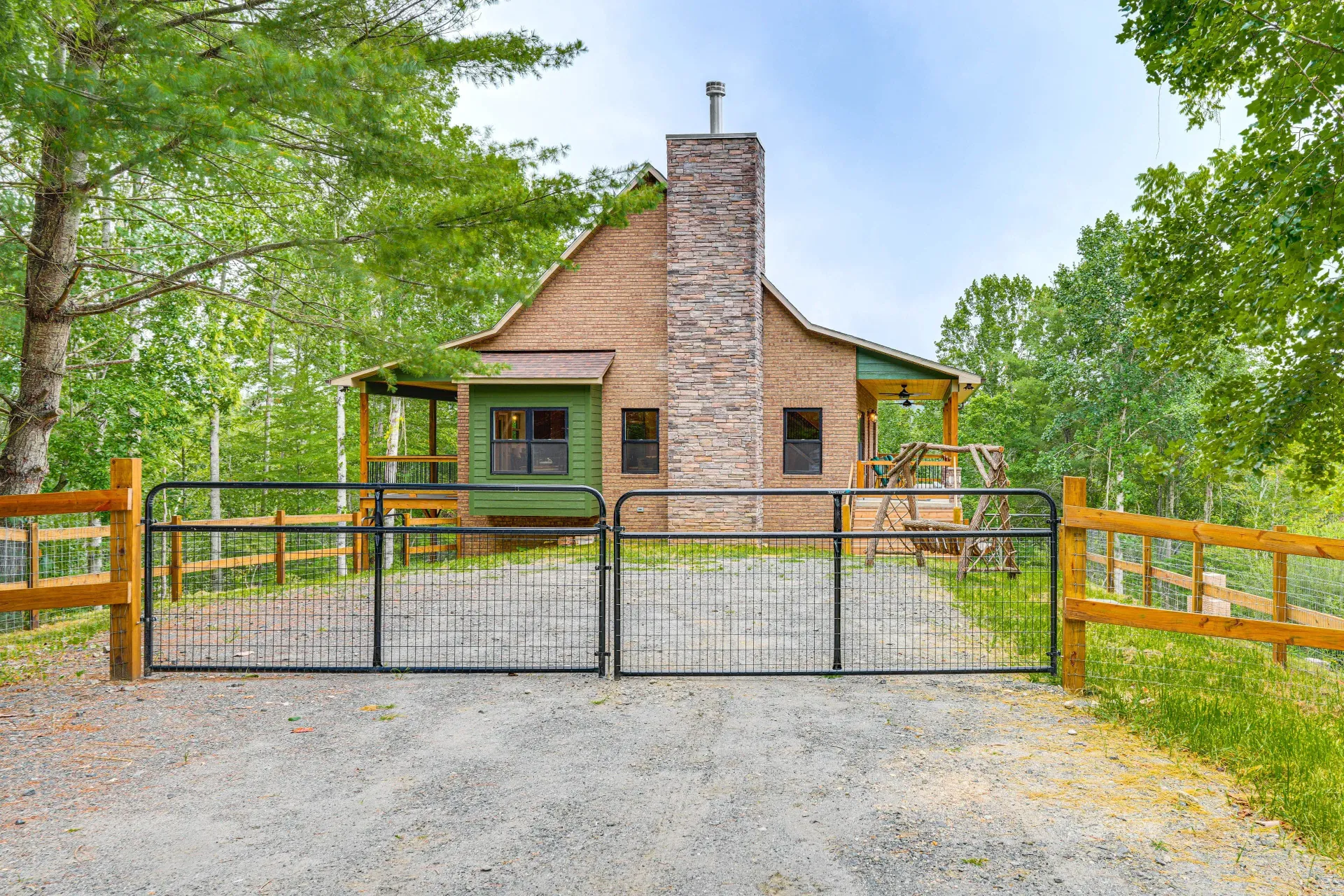 Stone cabin behind a black gate, framed by green trees and a gravel driveway