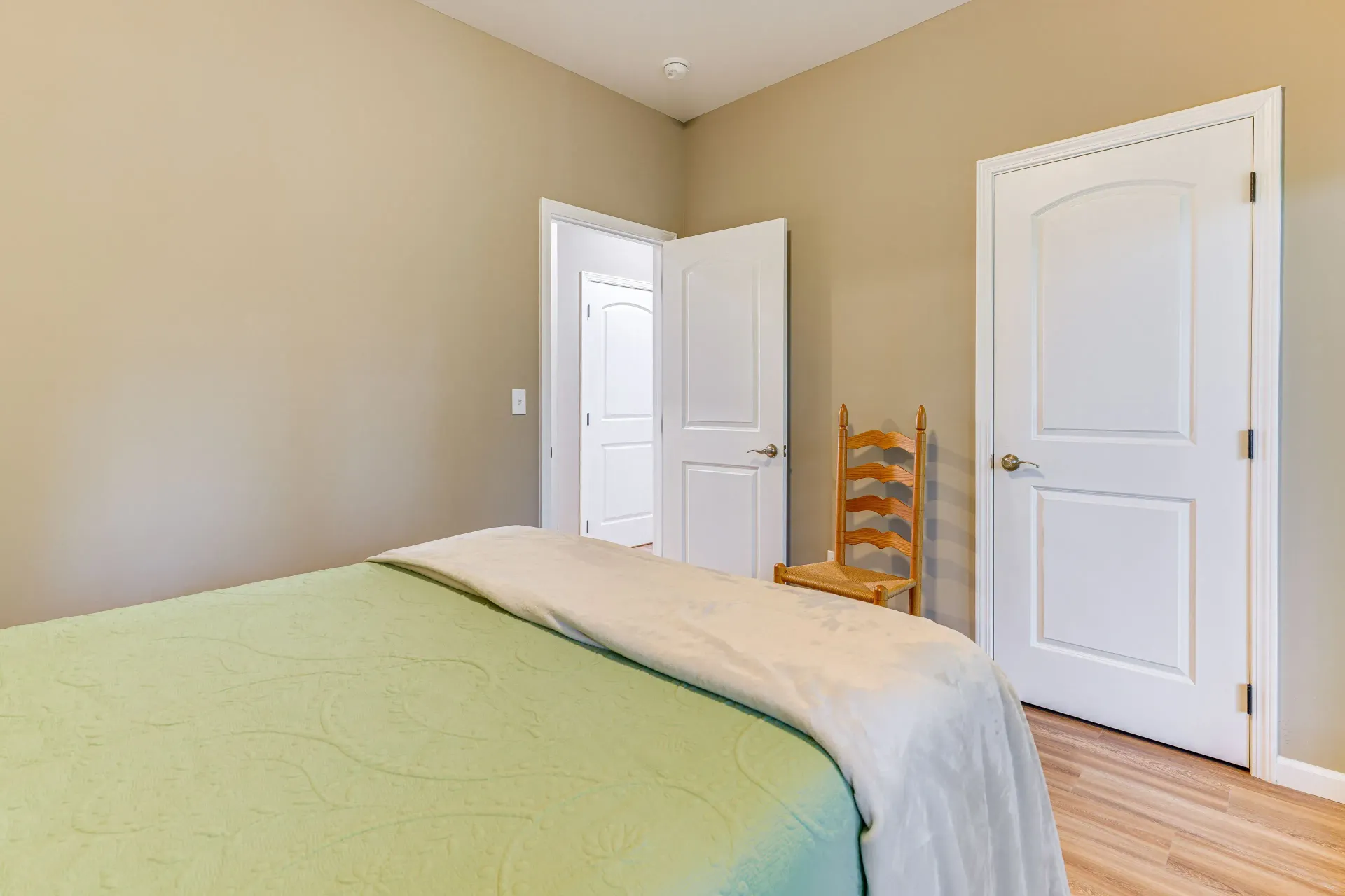 Empty beige bedroom with white doors, light wood floor, and a green bedspread.