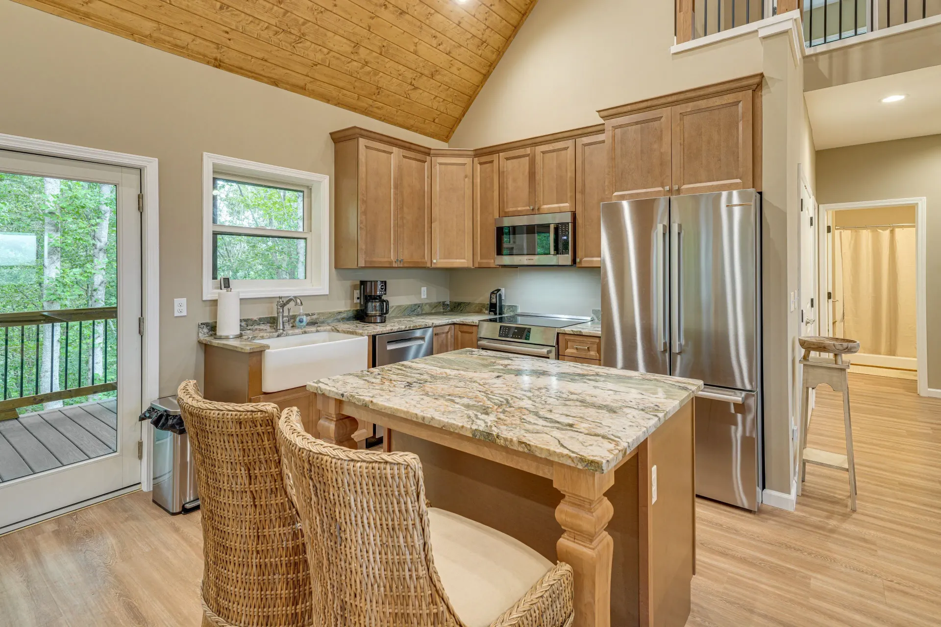 Bright kitchen with granite island, wicker chairs, stainless-steel refrigerator, and vaulted wood ceiling.