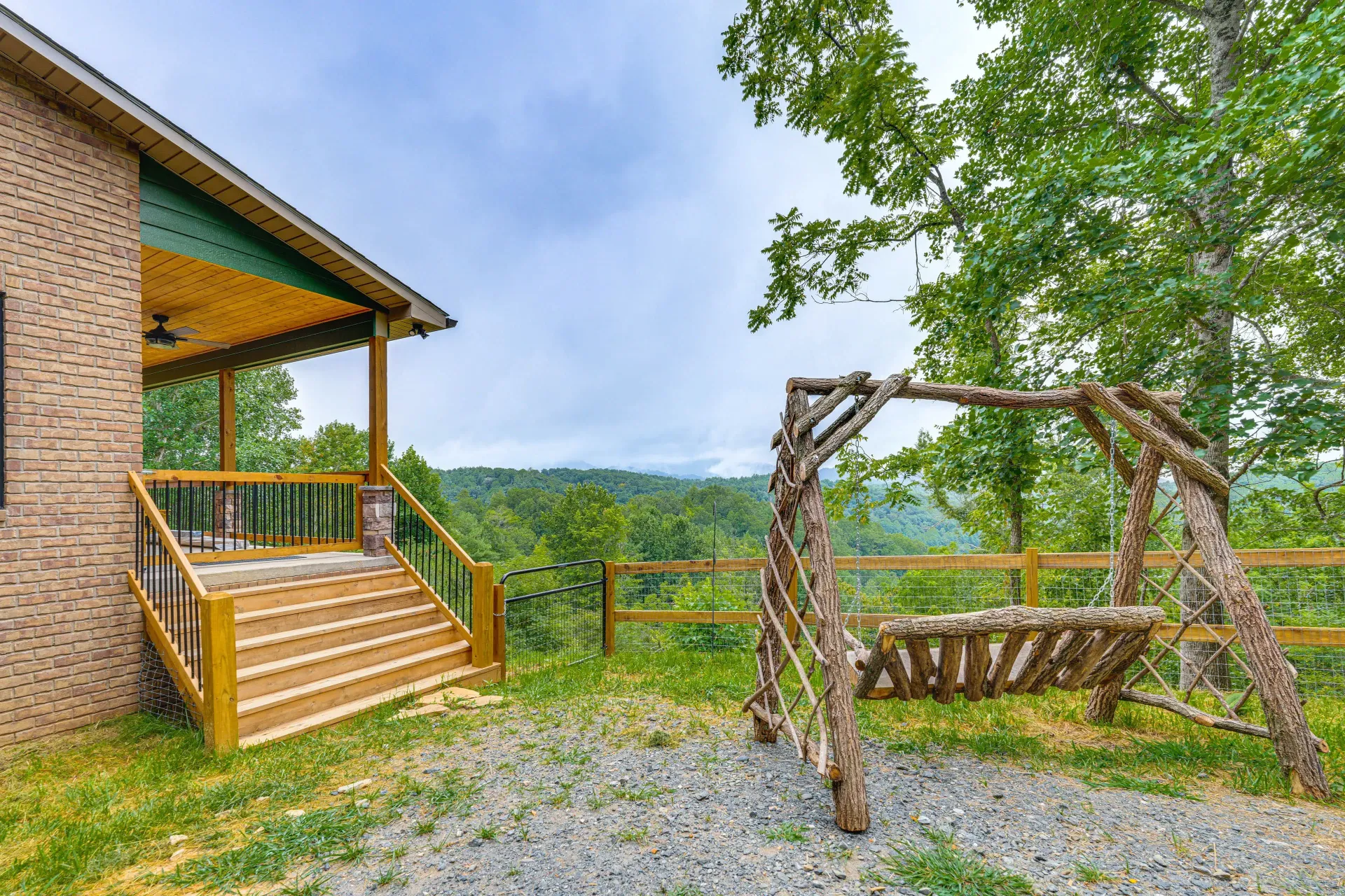 Brick house deck with wooden stairs overlooking a forested valley, beside a rustic swing on gravel.