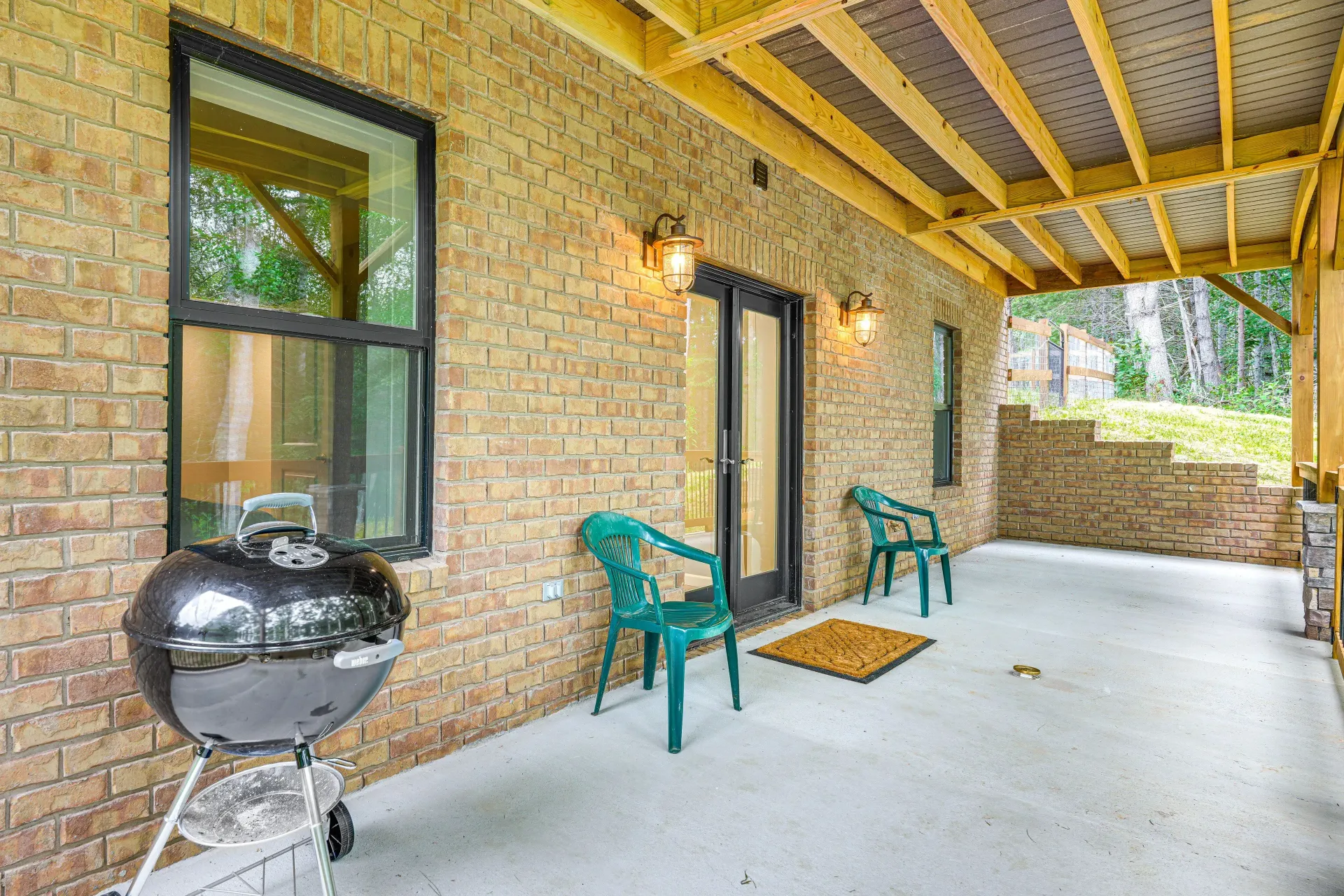 Covered brick patio with grill, two green chairs, and a wall light under a wood ceiling.