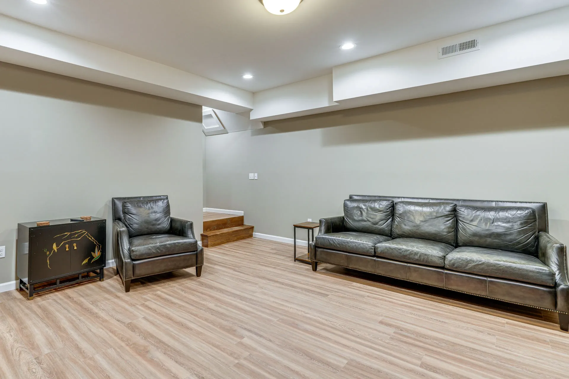 Basement lounge with gray leather sofa, chair, and small decorative cabinet on wood floors