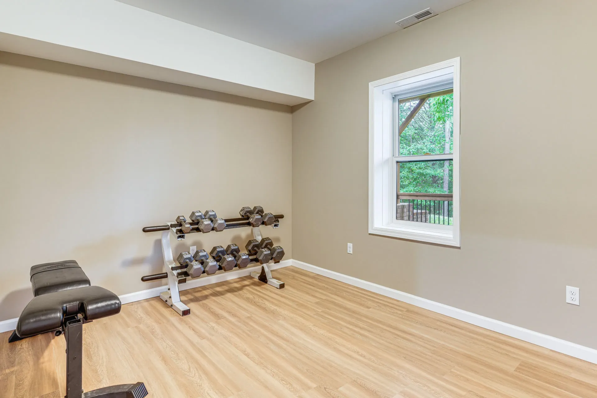 Empty home gym with hardwood floor, bench, dumbbells, and a window overlooking greenery