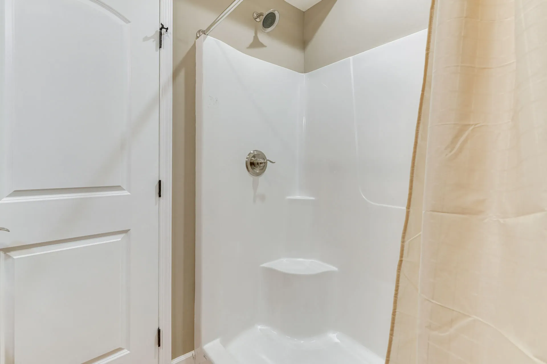 White shower stall with built-in shelves and beige curtain beside a white cabinet door