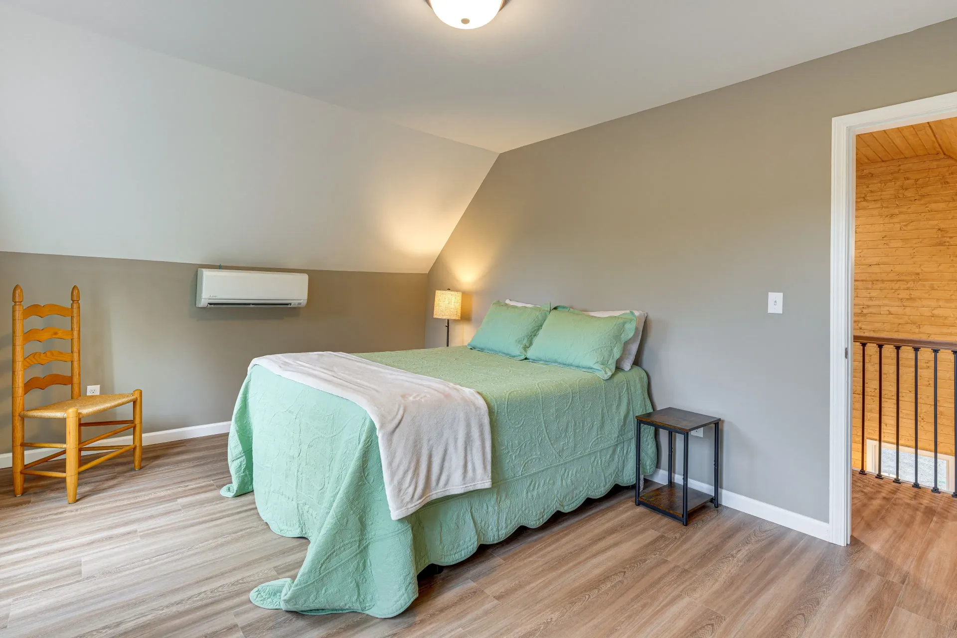 Spacious attic bedroom with green bedspread, wooden chair, and sloped white ceiling