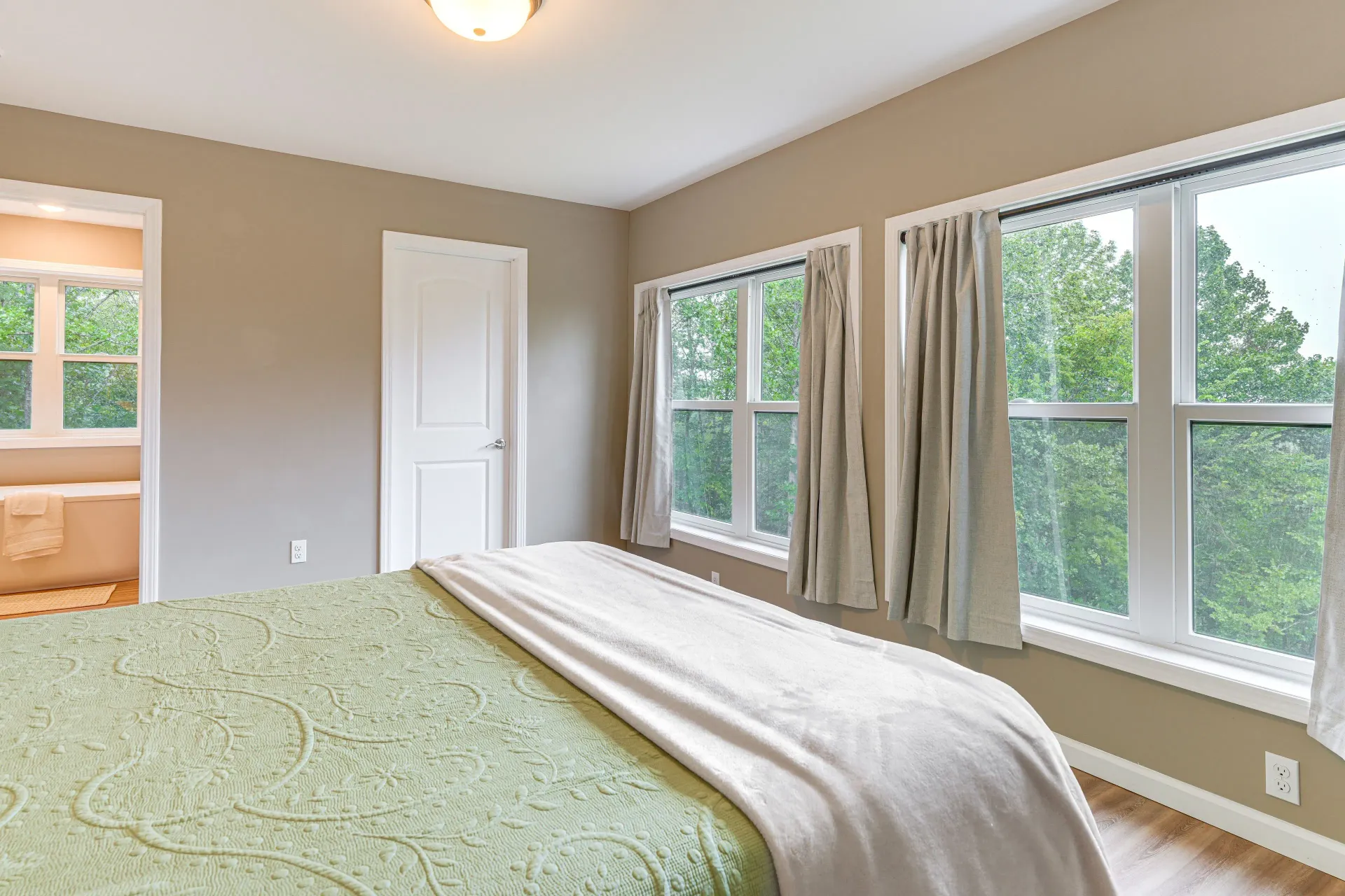 Bright bedroom with green bedspread, large windows, and a small bathroom visible through the open door.
