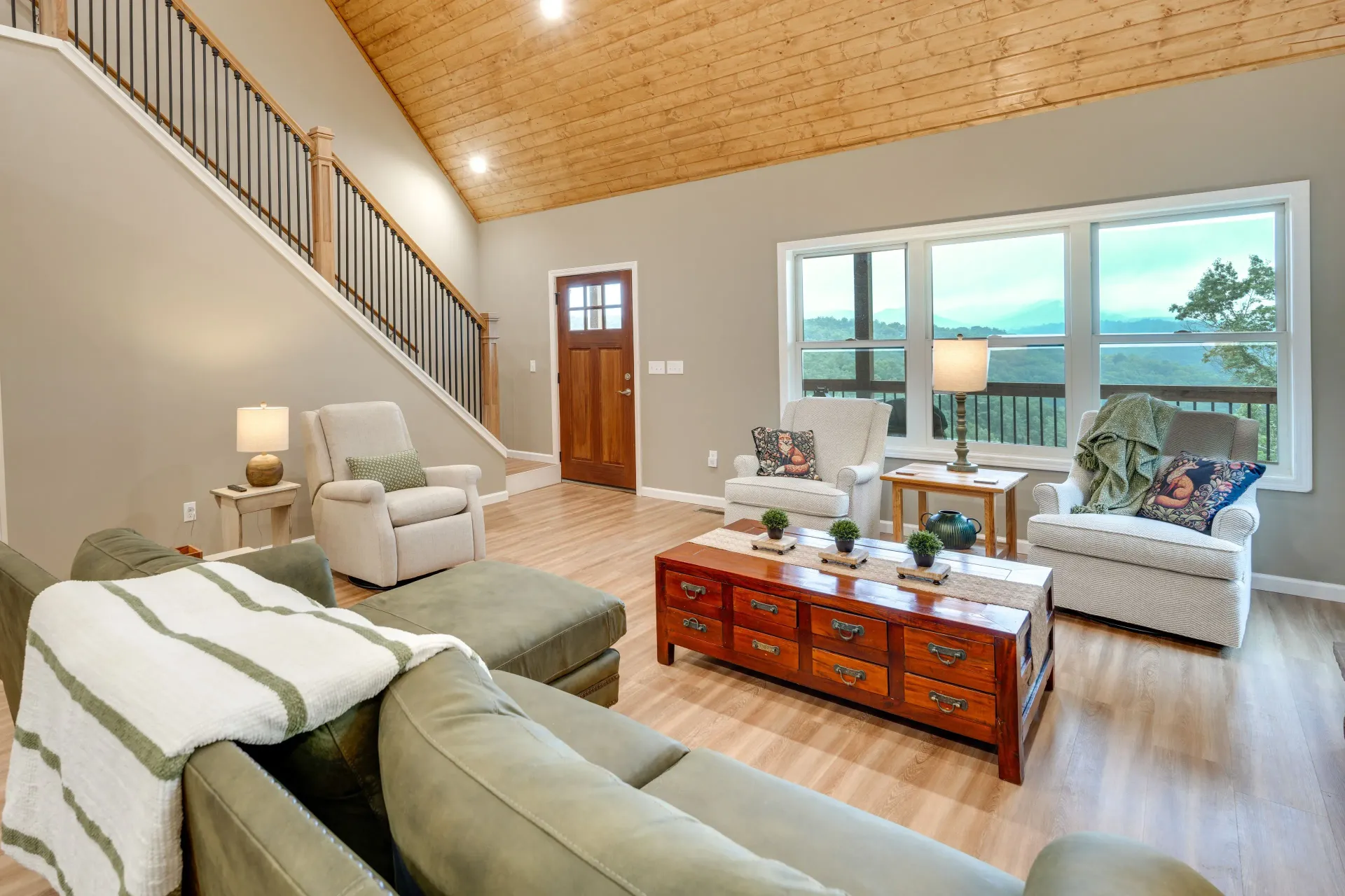 Bright living room with beige sofas, wood coffee table, staircase, and large windows overlooking a deck
