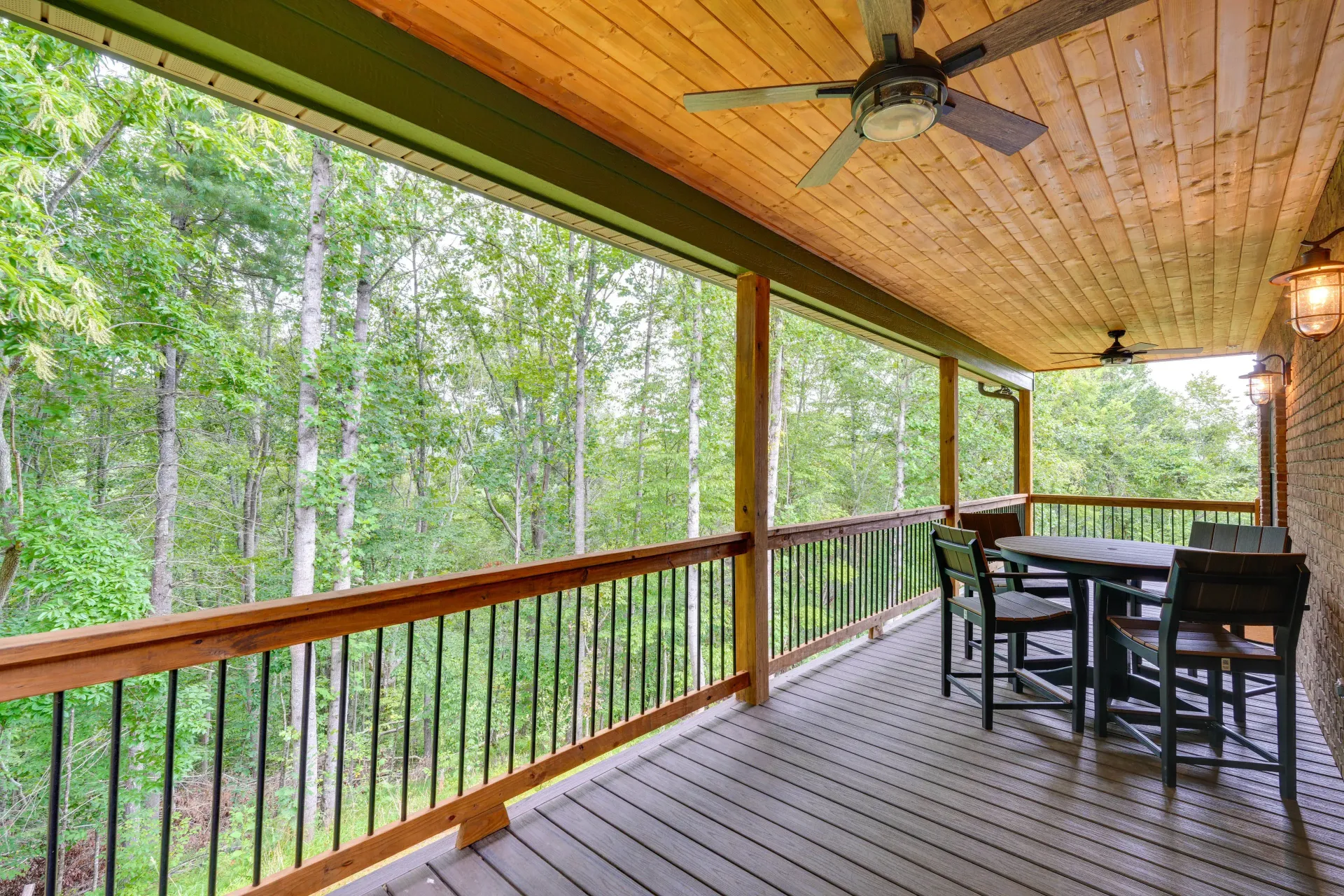 Covered wooden porch with screened railings, ceiling fan, and outdoor dining table overlooking a wooded area