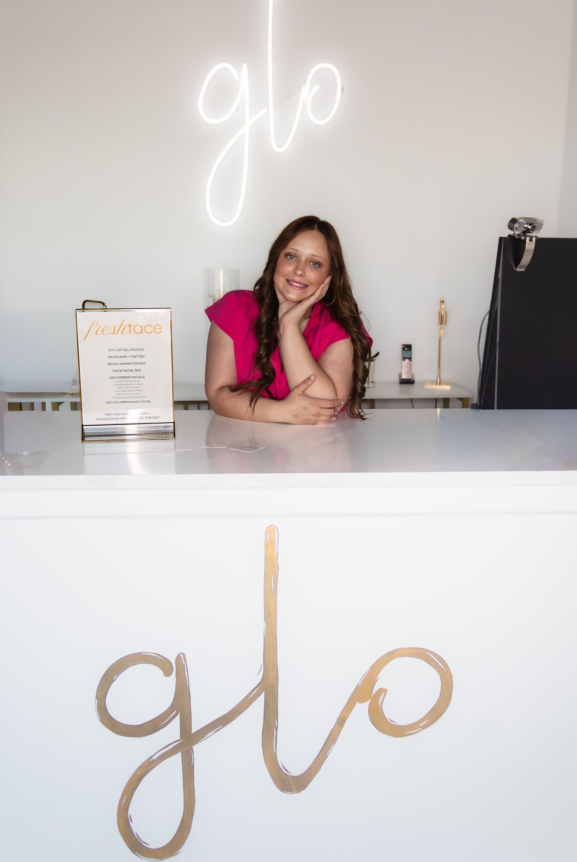 Josi in a pink top smiling behind a white counter in a bright shop with a glowing “glo” sign.