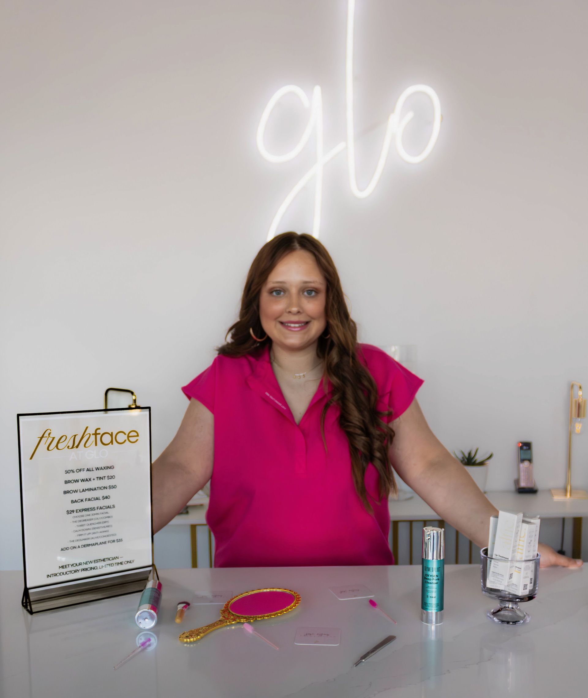 Woman in a bright pink blouse stands behind a table with jewelry and display items under a neon sign.