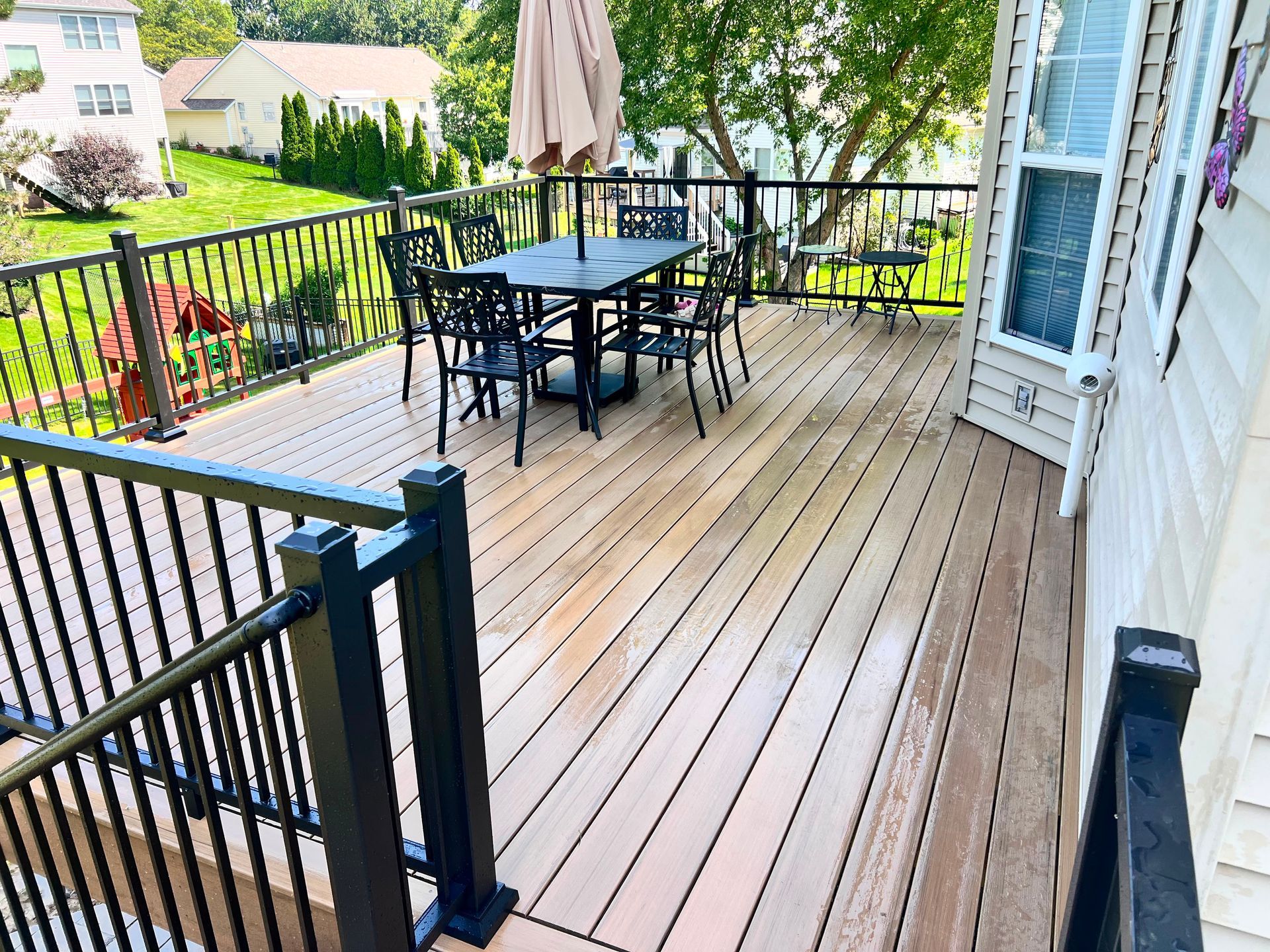 Spacious wooden deck with black patio table and chairs, railing, and a closed umbrella near a house.
