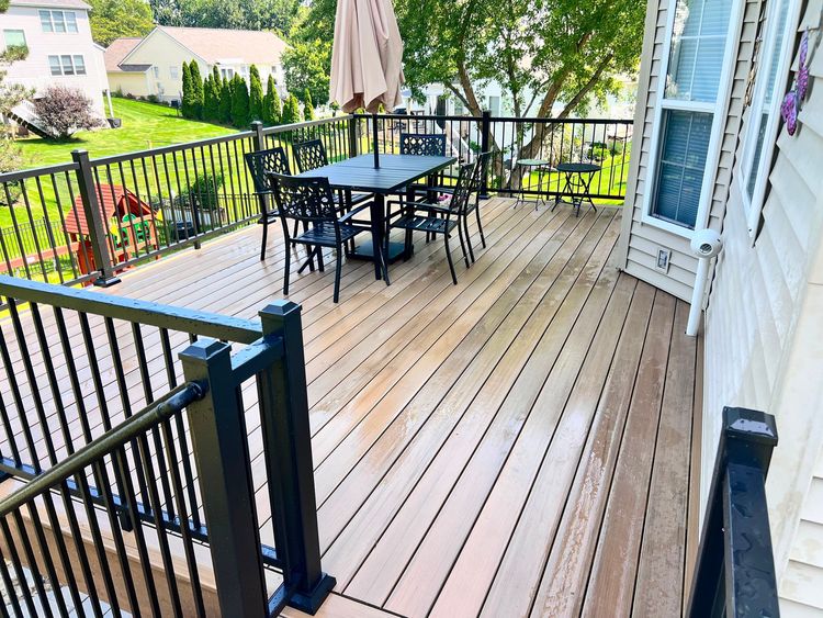 Spacious wooden deck with black patio table and chairs, railing, and a closed umbrella near a house.