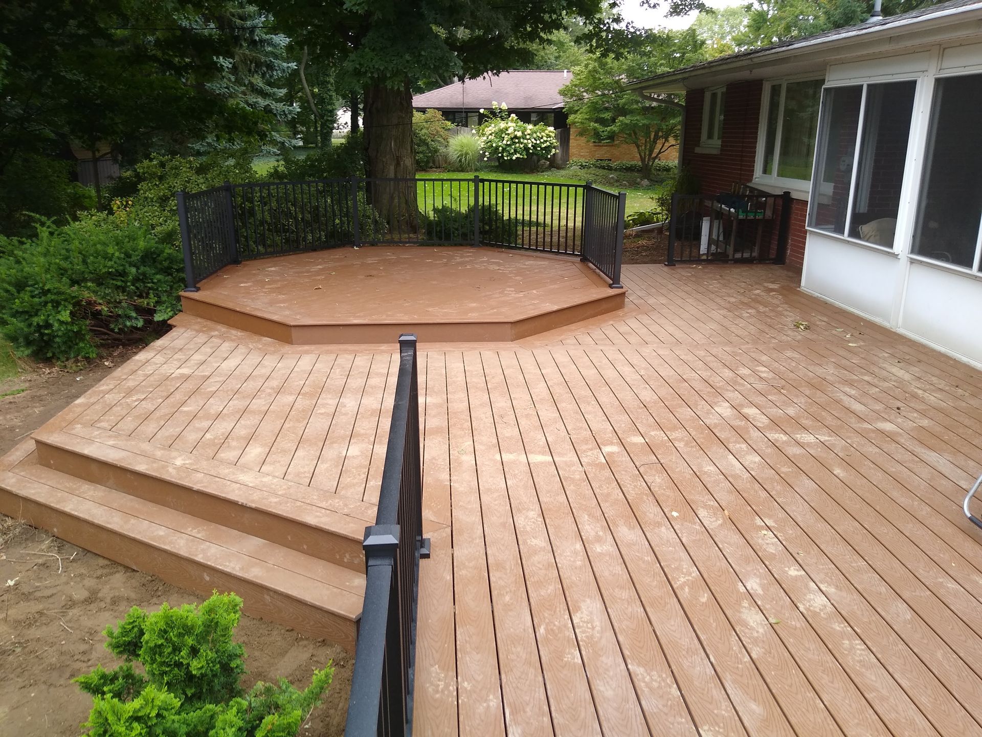 Wooden deck with an octagonal raised platform, black railing, and a house patio surrounded by greenery