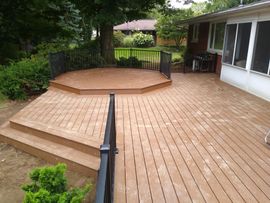 Wooden deck with a raised octagonal platform, steps, and black railing beside a house and trees.