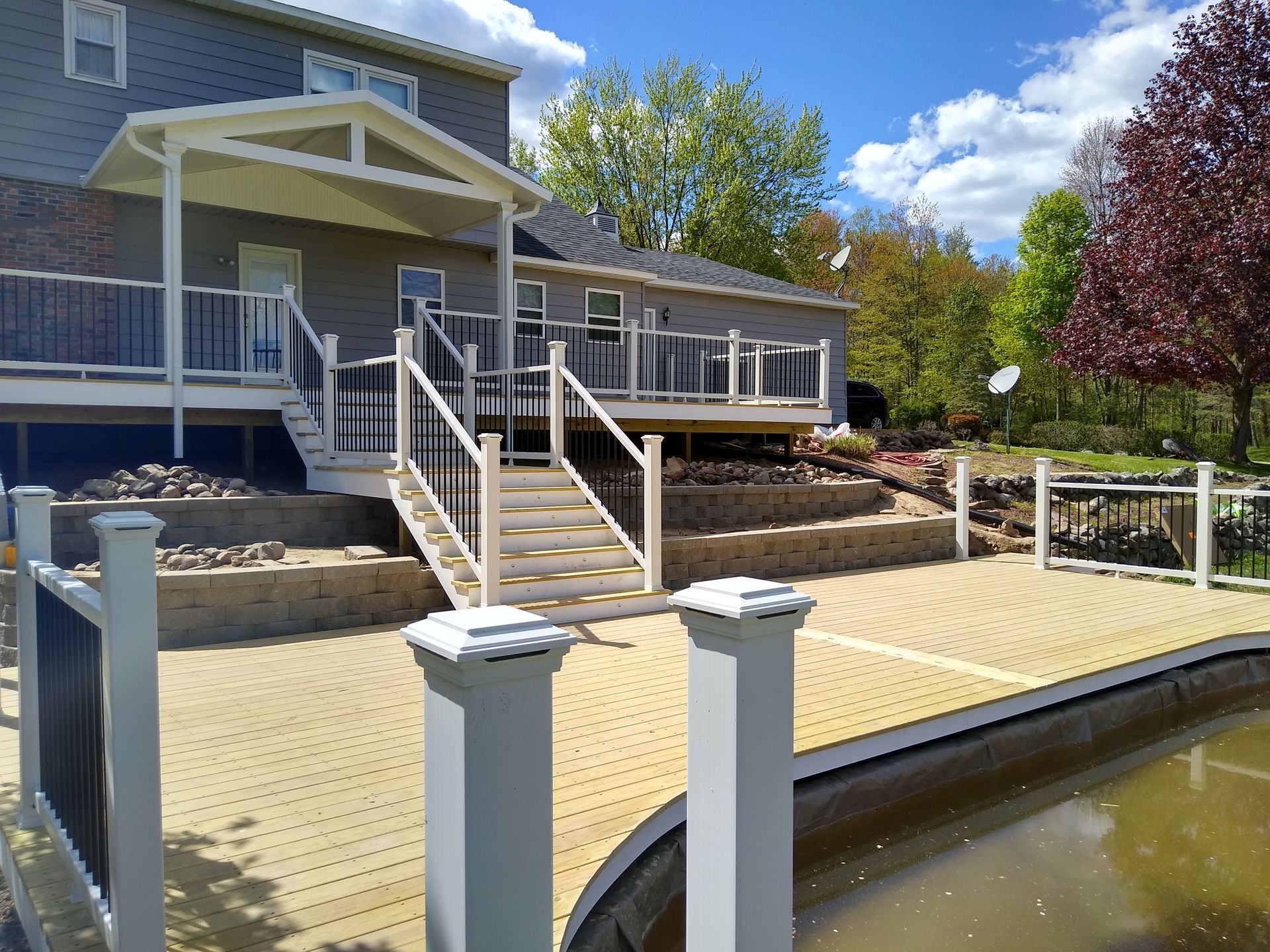 Backyard deck with stairs, stone retaining walls, and a pool beside a house on a sunny day