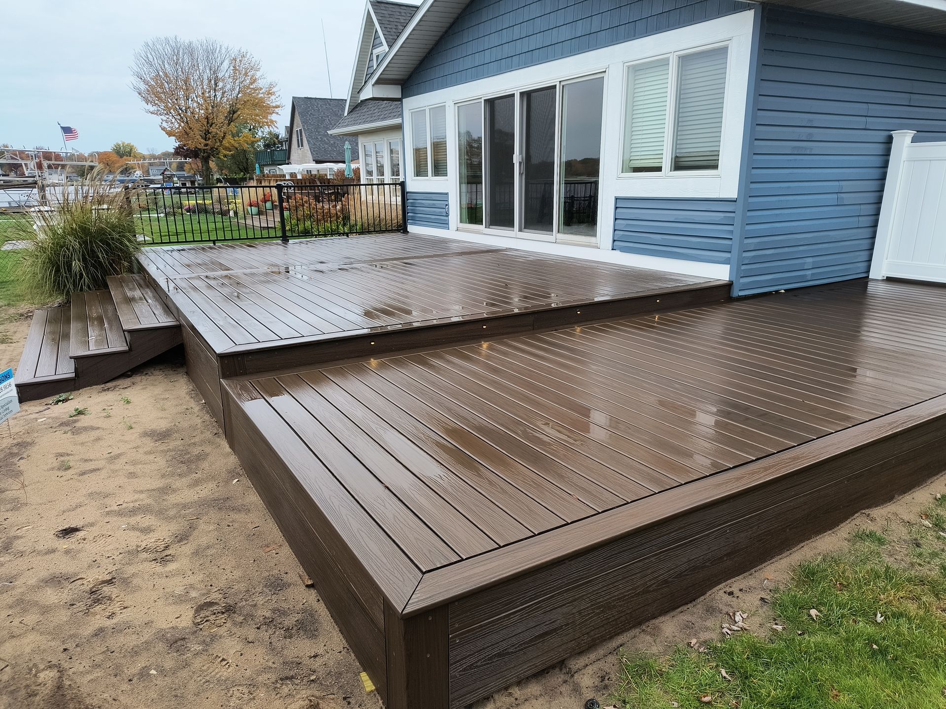 Blue house with large windows and wet wooden deck beside a sandy yard by the water