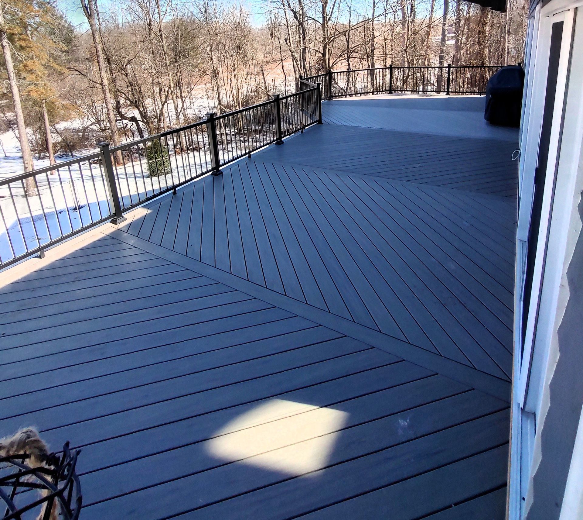 Empty gray composite deck with black railing overlooking snowy trees on a sunny day