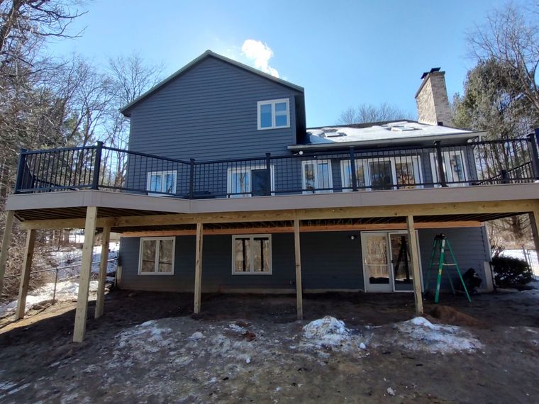 Two-story gray house with a deck, black railing, and a snowy yard under a blue sky