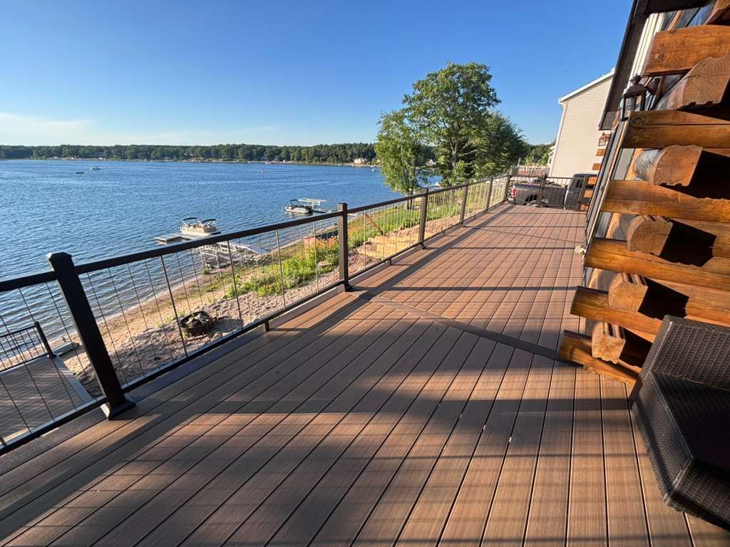 Wooden boardwalk beside a calm lake, with railings, rocks, and trees in sunlight