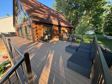 Wooden deck with black wicker lounge chairs beside a rustic cabin, surrounded by trees and sunlight.