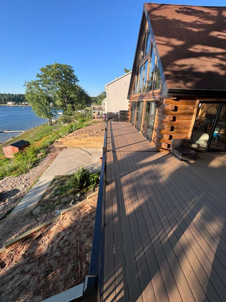 Wooden deck beside a lake, with a log cabin and bright blue sky at sunset.