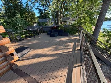 Wooden deck overlooking a lake, with black railings, trees, and outdoor seating in the shade.