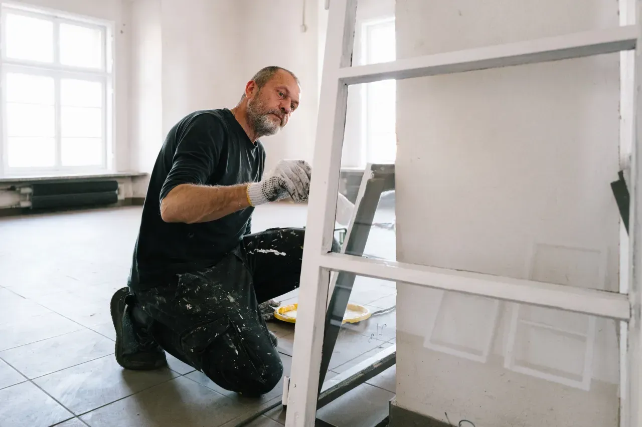 Painter kneeling beside a white ladder, painting a room with white walls and floors.