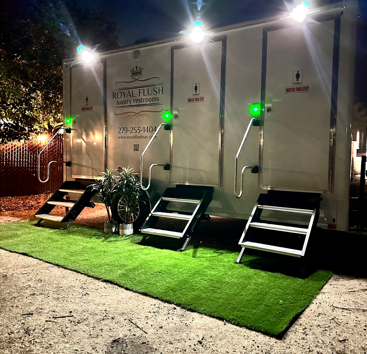 Illuminated mobile restroom trailers at night with three steps and green artificial turf